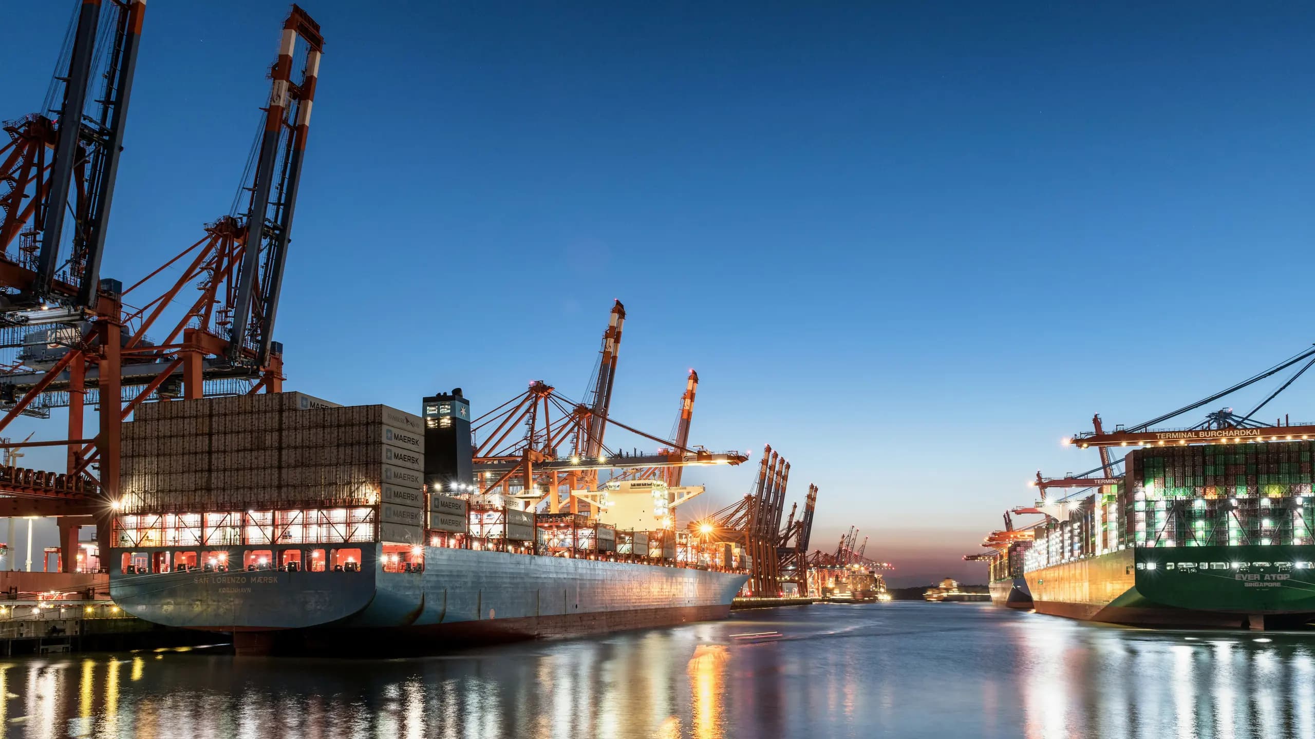 Container ships berthed at a terminal at blue hour, cranes lit against a deep blue sky